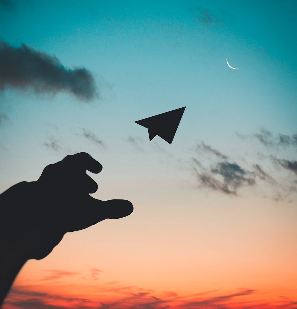 A silhouette of a hand launching a paper plane against a colorful dusk sky with a crescent moon.